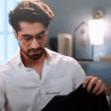 a man with a beard wearing glasses and a white shirt is holding a black shirt .