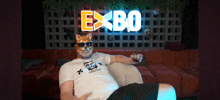 a man wearing a mask and sunglasses sits in front of a sign that says expo