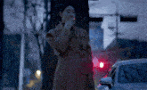 a woman in a brown coat is smoking a cigarette on the sidewalk