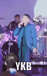 a man in a blue jacket is singing into a microphone on a stage with a drummer in the background .
