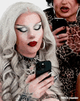 two drag queens are looking at their cell phones and one has a tattoo on her arm