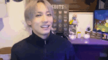 a young man is smiling in front of a sign that says shots