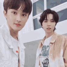 two young men are standing next to each other in front of a window and looking at the camera .