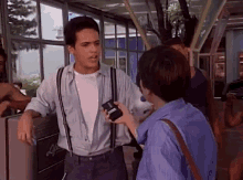 a man in suspenders is talking to a woman holding a camera