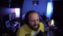 a man with a beard wearing headphones and a yellow shirt with the letter e on it stands in front of a microphone