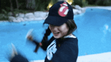 a woman wearing a rolling stones hat stands in front of a blue pool