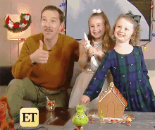 a man is giving a thumbs up while two little girls are playing with a gingerbread house