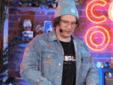 a man wearing a beanie and a denim jacket stands in front of a neon sign that says co.