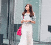 a woman in a blue shirt and white pants is standing in front of a glass door holding a pink purse .