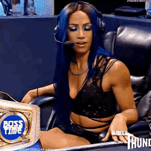a woman with blue hair is sitting in a chair wearing headphones and holding a wrestling championship belt .