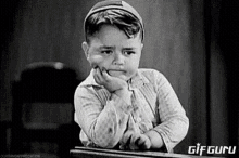 a black and white photo of a little boy sitting at a desk with his hand on his chin .