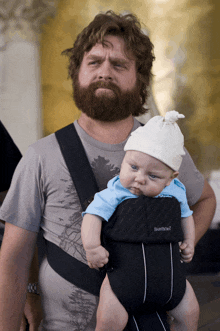 a man with a beard is carrying a baby in a carrier that says babybjorn