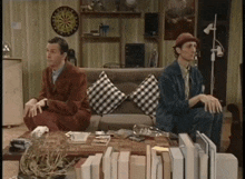 two men are sitting on a couch with a dart board on the wall behind them