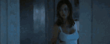 a woman in a white tank top is standing in a dark room and looking at her phone .