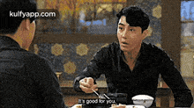 two men are sitting at a table eating food and one of them is talking to the other .