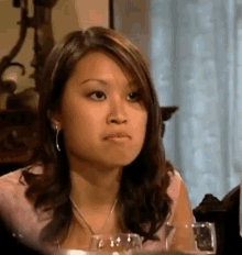 a woman is sitting at a table with a glass of wine and making a face .