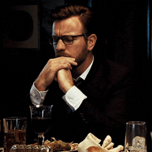 a man wearing glasses sits at a table with a plate of food and a glass of wine