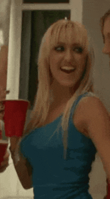 a blonde woman in a blue tank top is holding two red cups and smiling .