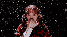 a girl in a red plaid poncho is standing in the snow blowing snow from her mouth .