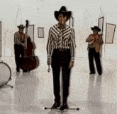 a man in a cowboy hat is singing into a microphone while two men play instruments behind him .