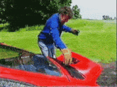 a man in a blue shirt is standing next to a red car in a field