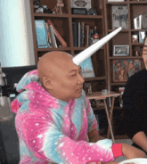 a bald man wearing a pink and blue unicorn pajama top has a paper horn on his head