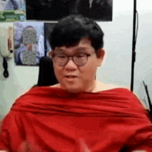 a man wearing glasses and a red shirt is sitting in front of a white wall .