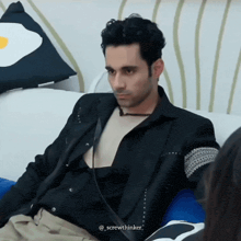 a man in a black jacket is sitting on a couch with an egg pillow in the background