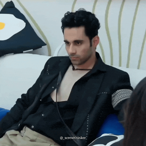 a man in a black jacket is sitting on a couch with an egg pillow in the background