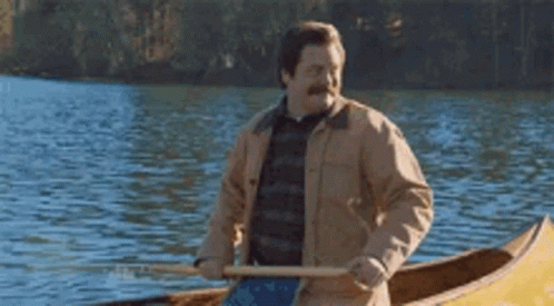 Ron from Parks and Rec, a white man with a great mustache and a canvas jacket in a yellow canoe paddling and smiling