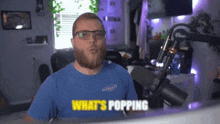 a man with a beard wearing glasses and a blue shirt says what 's popping in front of a microphone