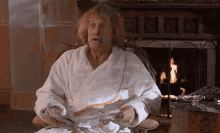 a man in a bathrobe is sitting in front of a fireplace smoking a cigarette and reading a newspaper .