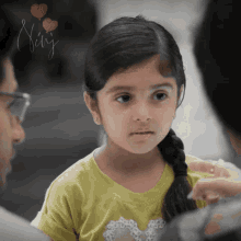 a little girl in a yellow shirt with a heart on it looks at the camera with a sign above her that says " tiny "