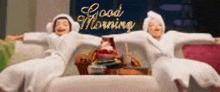two women in bathrobes are sitting on a bed with their arms outstretched and the words good morning written in the background