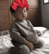 a baby is sitting on a bed with his eyes closed and has a red hair sticker on his head .