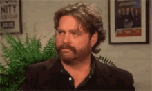 a man with a beard and long hair is sitting in front of a wall with a picture on it .