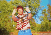 a man in an american flag shirt is giving the middle finger while standing in front of trees .