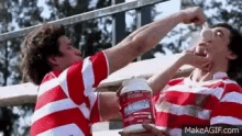two men in red and white striped shirts are drinking from a bottle of protein powder .