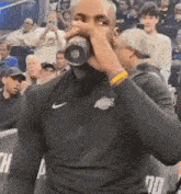 a man in a black nike shirt is drinking from a bottle while standing in front of a crowd .