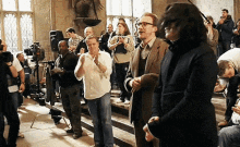 a group of people are standing in a room and one of them is wearing glasses and a black coat
