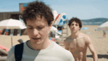 two shirtless men are walking on a beach with a lifeguard ladder in the background