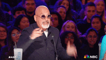 a bald man wearing sunglasses and a suit stands in front of a crowd with a cup that says nbc on it