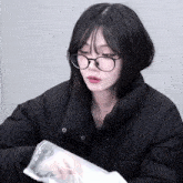 a woman wearing glasses and a black jacket holds a plastic bag