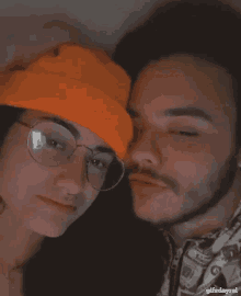 a man and a woman are sticking their tongues out . the woman is wearing glasses and an orange hat .