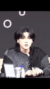 a young man with long black hair is sitting at a table with a bottle of water