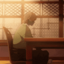a man in a green shirt sits at a wooden table in front of a window
