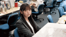 a woman in a suit sits at a desk in an office looking at the camera