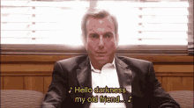 a man in a suit is sitting on a couch and singing hello darkness my old friend .