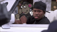 a man in a black hat is sitting in a car with a cartoon rat behind him