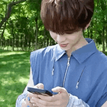 a young man in a blue shirt is holding a cell phone in his hands in a park .